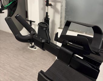 May include: A black bicycle triathlon handlebar extension set featuring textured grips, a central mounting bracket for a cycling computer, and a bottle cage attached to the side. The assembly includes a padded arm rest with a grid pattern surface.