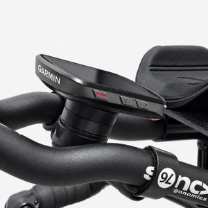May include: A black Garmin cycling computer mounted on black bicycle handlebars. The device features a dark screen, side buttons with a red accent, and a mounting stem. The handlebars include black padded arm rests and a label reading sync ergonomics.