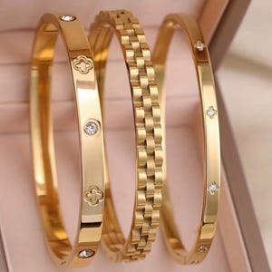 May include: A set of three gold-toned metal bangles. The first bangle features alternating clover-shaped cutouts and small round clear stones. The second bangle has a textured, linked chain-style design. The third bangle is smooth with small, evenly spaced, four-pointed star-shaped clear stone settings.