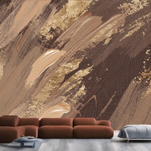 May include: Abstract art wallpaper with a textured design of brown, tan, and gold brushstrokes. The artwork has a modern aesthetic, suitable for interior design. The image shows a brown sectional sofa, a small round table, and a grey ottoman.