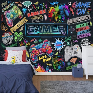 Könnte beinhalten: Eine schwarze Wandtapete mit farbenfrohen Grafiken zum Thema Videospiele, darunter Controller und Texte wie "Gamer", "Fortnite" und "Minecraft". Die Szene umfasst ein Bett mit einer blauen Bettdecke, Kissen und einen kleinen Nachttisch.