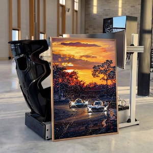 Könnte beinhalten: Gerahmtes Gemälde, das einen Sonnenuntergang über einer Rennstrecke mit zwei Autos zeigt. Das Kunstwerk zeigt lebendige Orange-, Gelb- und Rottöne am Himmel, mit Bäumen und den Rennwagen im Vordergrund. Links ein schwarzer Rennsitz.