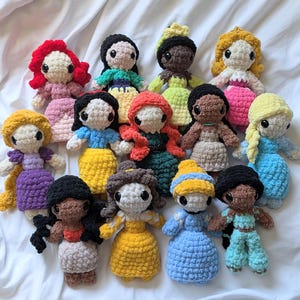 Puede incluir: Una colección de muñecas de ganchillo hechas a mano, cada una representando un personaje de princesa diferente. Las muñecas presentan varios colores de cabello y atuendos, incluyendo vestidos y accesorios. Las muñecas están dispuestas sobre un fondo de tela blanca.