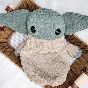 May include: A green and beige crocheted plush toy in the shape of a character with large ears and big eyes. The toy is sitting in a woven basket.