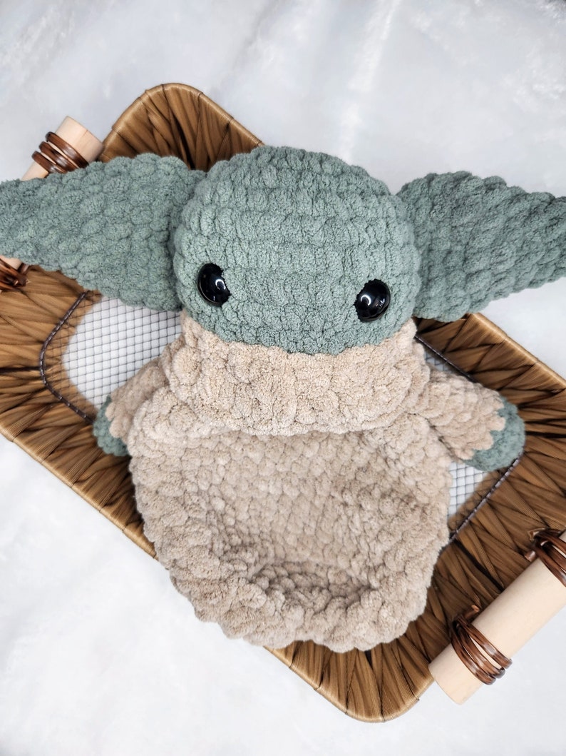 May include: A green and beige crocheted plush toy in the shape of a character with large ears and big eyes. The toy is sitting in a woven basket.