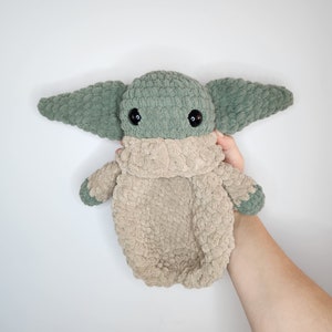 May include: A green and beige crocheted plush toy resembling a popular fictional character. The toy has large ears and black eyes.