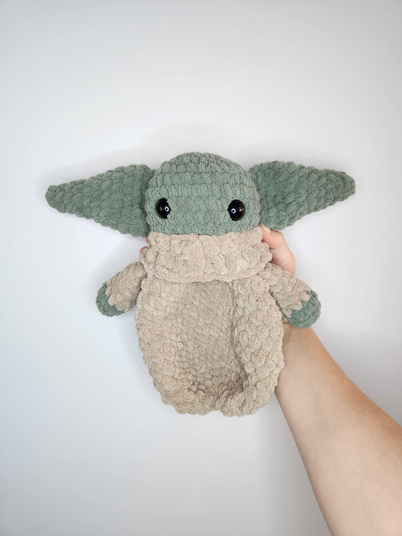 May include: A green and beige crocheted plush toy resembling a popular fictional character. The toy has large ears and black eyes.