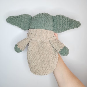 May include: A crocheted plush toy in the shape of a green alien character with large ears. The toy is wearing a beige crocheted coat.