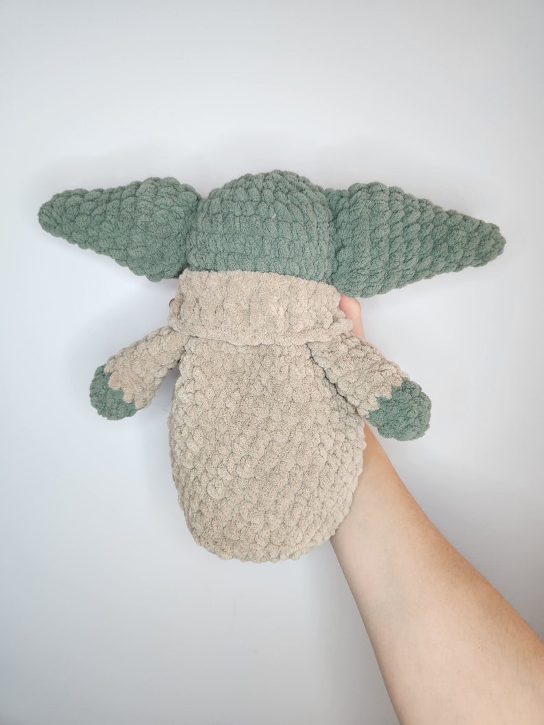 May include: A crocheted plush toy in the shape of a green alien character with large ears. The toy is wearing a beige crocheted coat.