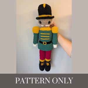 May include: A handmade crochet nutcracker doll in a classic uniform. The doll features a black top hat with yellow trim, a green jacket with yellow epaulets, red pants, and black boots. The text "PATTERN ONLY" is at the bottom.