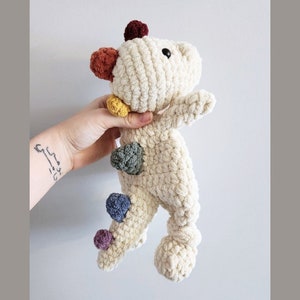 May include: A crocheted stuffed animal shaped like a dinosaur with a cream coloured body and colourful accents. The dinosaur has a long tail with a pom-pom at the end.