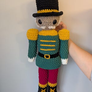 May include: Crocheted nutcracker doll in a traditional uniform. The doll features a black hat with yellow trim, a green jacket with gold accents, red pants, and black boots. The doll is approximately 12 inches tall.