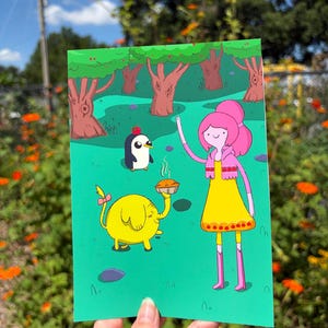 May include: A rectangular art print featuring a cartoon scene with a pink-haired character in a yellow dress, a yellow elephant with a pink bow, and a small penguin with a red apple on its head. The characters stand in a green forest setting.