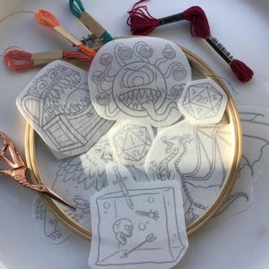 May include: A set of embroidery patterns featuring various fantasy creatures and dice. The patterns are printed on white fabric and are ready to be stitched. The patterns include a dragon, a monster with multiple eyes, a chest, a bird, and a dice with the number 20 on it.