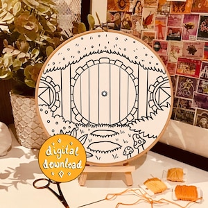 Può includere: Un disegno in bianco e nero di una porta rotonda con una finestra rotonda al centro, circondata da un arco in pietra e un'area erbosa con funghi. Il disegno è su una tela circolare bianca, che è montata su un supporto in legno. Le parole "download digitale" sono scritte in giallo su uno sfondo a forma di diamante.