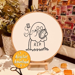 May include: A black and white embroidery pattern of a ghost holding a pie with the text "R.I.P." on a tombstone. The pattern is on a white embroidery hoop.