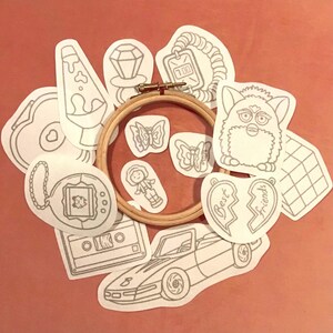 May include: A selection of grey line art patches with various designs, such as a Furby, butterflies, a cassette tape, a car, and a broken heart with the words "Best Friends." The patches are arranged around a wooden embroidery hoop.