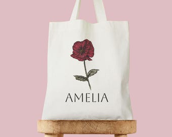 May include: White tote bag with a red flower illustration and the name Amelia printed in black.