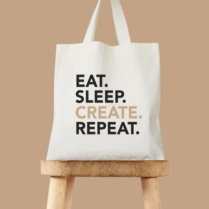 May include: White canvas tote bag with black text that reads "EAT. SLEEP. CREATE. REPEAT." The bag is sitting on a wooden stool.