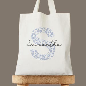 Könnte beinhalten: Weiße Einkaufstasche mit einem floralen Design in Form des Buchstabens "S" und dem Namen "Samantha" in Schwarz gedruckt.