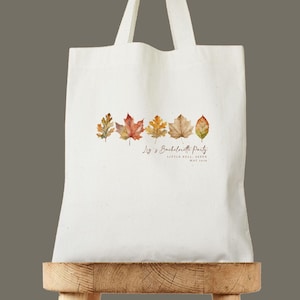 Könnte beinhalten: Eine weiße Tragetasche mit einem Aquarelldesign von Herbstblättern in Braun-, Orange- und Gelbtönen. Unter den Blättern ist der Text "Liz's Bachelorette Party" zusammen mit Ort und Datum aufgedruckt.