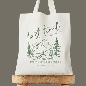 May include: Off-white canvas tote bag with a green illustration of mountains, trees, and a tent. Text reads "last trail before the veil" and "Julia's Bachelorette, Lake Tahoe, CA, 2022". The bag has two handles.