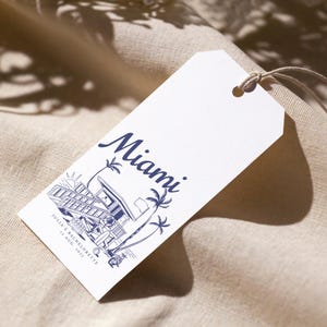May include: White tag with a navy blue illustration of a lifeguard station, palm trees, and the word "Miami." The text "JULIA'S BACHELORETTE 12 AUG. 2023" is also printed on the tag. A string is attached to the tag.