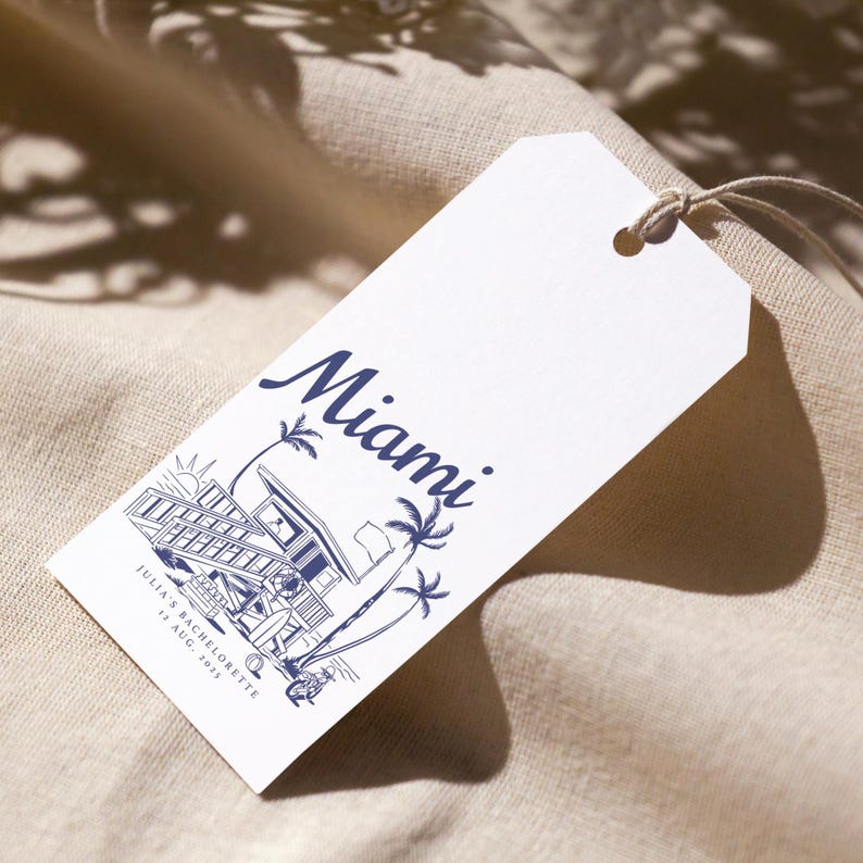 May include: White tag with a navy blue illustration of a lifeguard station, palm trees, and the word "Miami." The text "JULIA'S BACHELORETTE 12 AUG. 2023" is also printed on the tag. A string is attached to the tag.