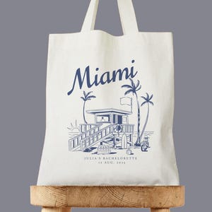 May include: Off-white tote bag with a blue illustration of a Miami lifeguard station, palm trees, and the word "Miami." The text "JULIA'S BACHELORETTE 12 AUG. 2023" is also printed on the bag. The bag has two white handles.