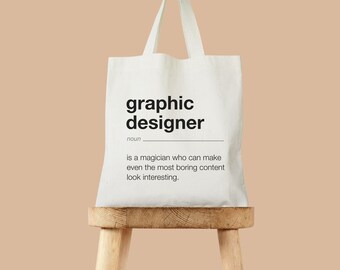 May include: White tote bag with the text "graphic designer" and the definition "is a magician who can make even the most boring content look interesting."