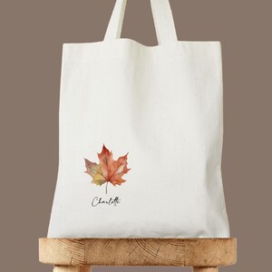 May include: A white tote bag with a watercolor maple leaf design in shades of red, orange, and yellow. The name "Charlotte" is written in a cursive font below the leaf. The bag has two handles and is displayed on a wooden stand.