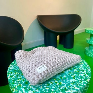 Puede incluir: Una almohada de crochet gris claro con una etiqueta blanca que dice "ALENNIGRAM" sobre una superficie moteada de verde y blanco.