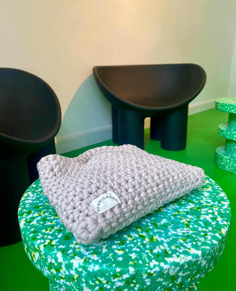 Puede incluir: Una almohada de crochet gris claro con una etiqueta blanca que dice "ALENNIGRAM" sobre una superficie moteada de verde y blanco.
