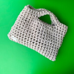 Puede incluir: Una bolsa de mano de crochet beige con un solo asa. La bolsa tiene una peque&ntilde;a etiqueta blanca con el texto "LENIC BRAND" en ella.