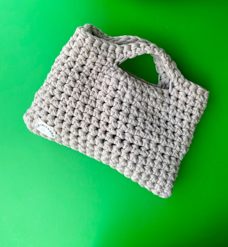 Puede incluir: Una bolsa de mano de crochet beige con un solo asa. La bolsa tiene una peque&ntilde;a etiqueta blanca con el texto "LENIC BRAND" en ella.