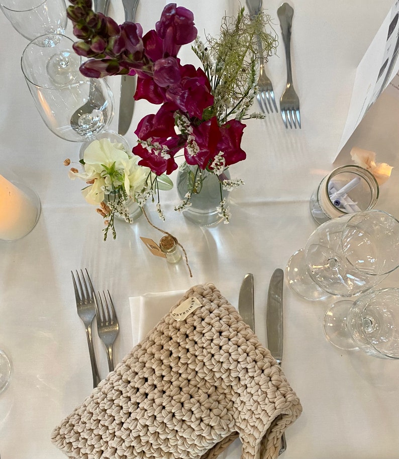 Puede incluir: Una bolsa de crochet beige con una peque&ntilde;a etiqueta que dice "hecho a mano". La bolsa est&aacute; sobre un mantel blanco con dos tenedores, un cuchillo y un vaso de agua. Tambi&eacute;n hay flores en un jarr&oacute;n y un peque&ntilde;o frasco de vidrio con una vela.