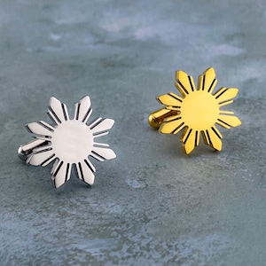May include: A pair of cufflinks, one silver and one gold, each with a sunburst design. The sunburst has a central circle with radiating lines. The cufflinks are presented on a textured, blue-grey surface. The design is reminiscent of a star.