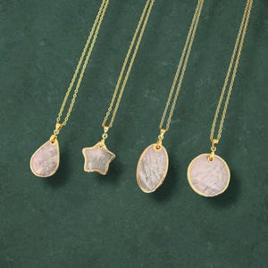 May include: Four gold necklaces with pendants of varying shapes: a teardrop, a star, an oval, and a circle. Each pendant features a pale pink stone with a gold-coloured border. The necklaces are presented against a dark green backdrop.