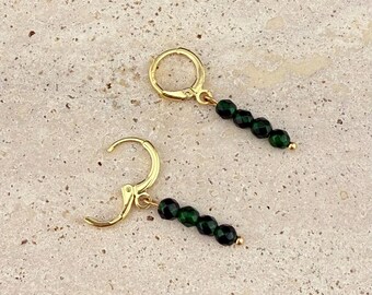 May include: A pair of gold-colored hoop earrings with a dangling row of five dark green beads. The earrings are displayed on a light beige, speckled surface.