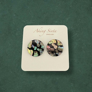 May include: Round stud earrings with a black base and iridescent abalone shell inlay. The earrings are presented on a white card with the text "Aking Sinta Jewellery". The background is a dark green. The earrings are a unique piece of jewellery.