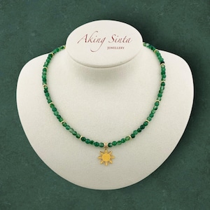May include: A green beaded necklace with a gold sun pendant. The necklace is displayed on a white bust against a dark green background. The brand name "Aking Sinta Jewellery" is printed at the top.