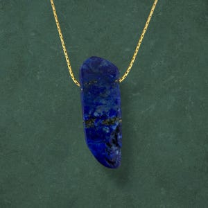 May include: A gold chain suspends a deep blue lapis lazuli pendant. The stone, with its irregular shape, features dark blue and gold inclusions. The necklace is set against a dark green backdrop, accentuating the pendant's colour and texture.