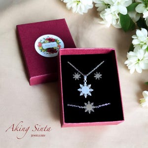 Peut inclure: Une boîte rouge contenant un collier, des boucles d'oreilles et un bracelet en argent. Le collier présente un motif de soleil blanc. La boîte est ouverte et les bijoux sont visibles. La boîte porte l'inscription "Aking Sinta Jewellery" en bas.