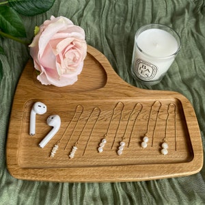 May include: A wooden tray with six pairs of gold chain earrings with pearl accents. The earrings are arranged in a row on the tray. There are two white wireless earbuds on the tray as well.