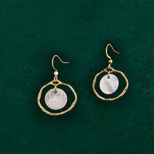 May include: A pair of gold-coloured earrings with a circular design. Each earring features a textured gold-tone outer ring and a white, circular pendant. The earrings hang from a simple hook. The background is a dark green colour.