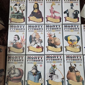 Pode incluir: Uma coleção de doze fitas VHS de Monty Python's Flying Circus dispostas em três filas. Cada estojo branco apresenta ilustrações surreais distintas, o título do programa em negrito e informações específicas sobre cada volume.