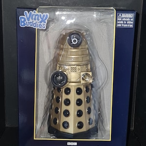 Könnte beinhalten: Goldfarbene Classic Dalek Vinyl-Figur aus der Doctor Who-Serie, verpackt in einer blauen Box. Die Figur hat schwarze Details und ist mit "Vinyl Buddies" gekennzeichnet. Die Box enthält einen Altershinweis.