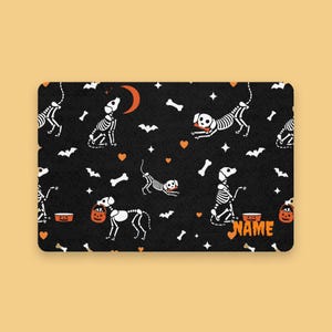 Könnte beinhalten: Eine rechteckige, schwarze Halloween-Matte mit einem sich wiederholenden Muster aus Skeletthunden, Knochen, Fledermäusen, Sternen und orangefarbenen Herzen. Die Matte hat einen Kürbis-Eimer und das Wort "NAME" in Orange.