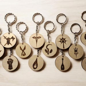 May include: Twelve wooden keychains with different designs, each with a silver key ring. The designs include a sword and shield, a hand with a swirl, a fist, a pair of crossed swords, a flame, a lute, an eye with a swirl, a crescent moon with leaves, a staff with a star, a helmet with horns, and a pair of crossed swords with a paw print.