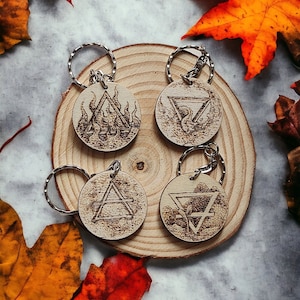 Peut inclure: Quatre porte-clés en bois avec des anneaux argentés, chacun gravé d'un symbole élémentaire différent : feu, air, terre et eau. Les porte-clés sont disposés sur une tranche de bois, entourés de feuilles d'automne colorées.